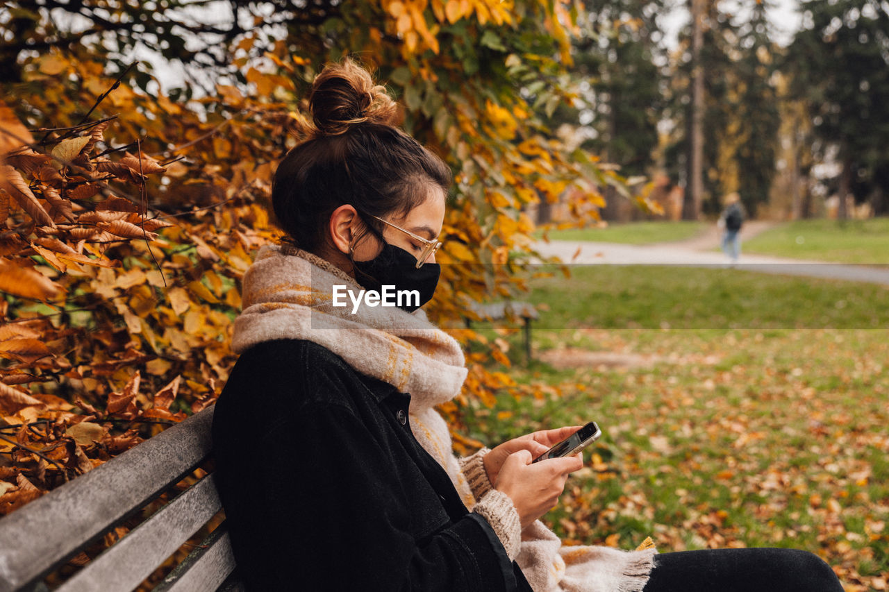 Woman wearing mask using smart phone sitting on bench