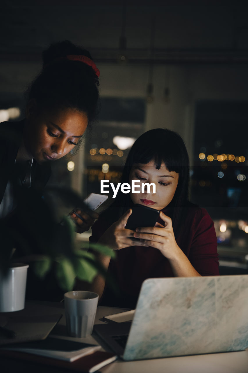 Confident female colleagues using smart phones while working late at coworking space