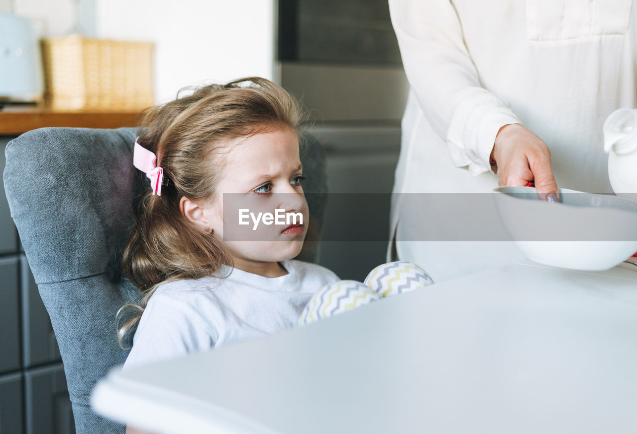Unhappy girl with long hair in home clothes with yogurt sitting at dinning table in kitchen at home