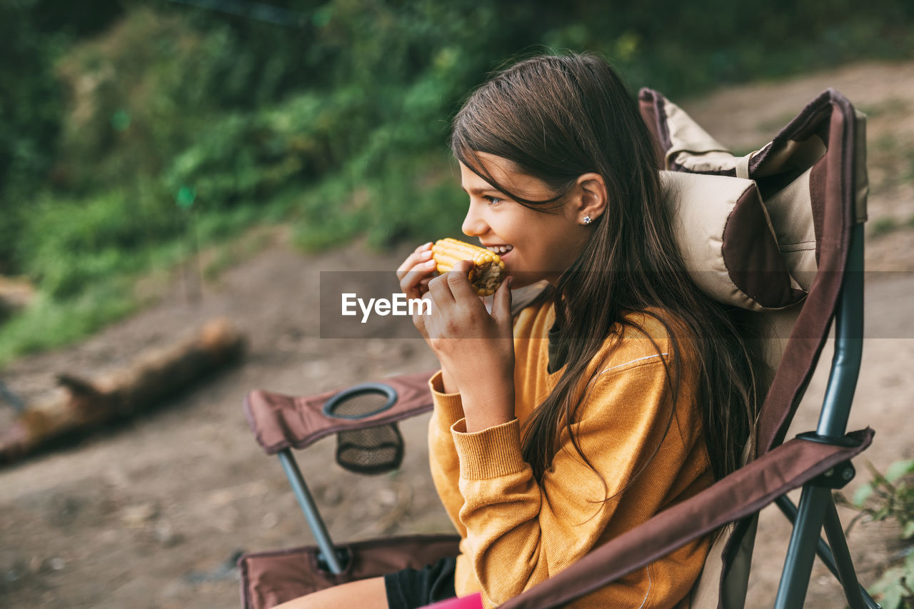 A teenage girl in a bright yellow sweater is sitting in a camping chair on the shore of the lake