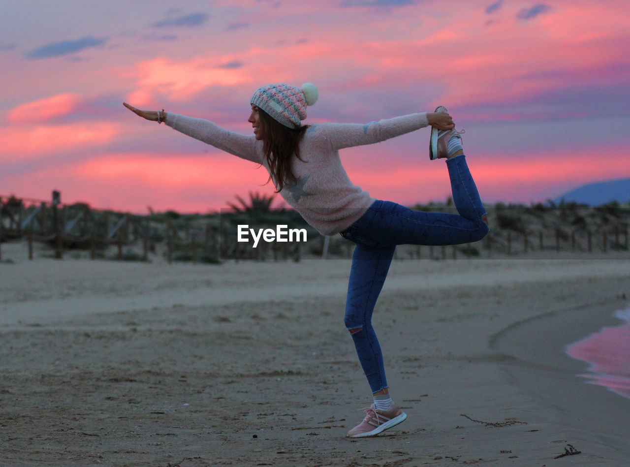 Woman doing yoga at beach against sky during sunset