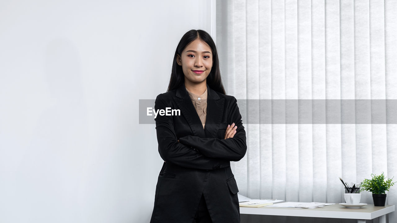 adult, one person, business, women, businesswoman, indoors, portrait, young adult, black, office, standing, formal wear, clothing, female, looking at camera, corporate business, outerwear, copy space, waist up, business finance and industry, arms crossed, front view, table, white-collar worker, smiling, photo shoot, long hair, black hair, hairstyle, emotion, businesswear, occupation, fashion, three quarter length, person, sitting, serious, sleeve, houseplant, furniture, collar, looking, contemplation, working