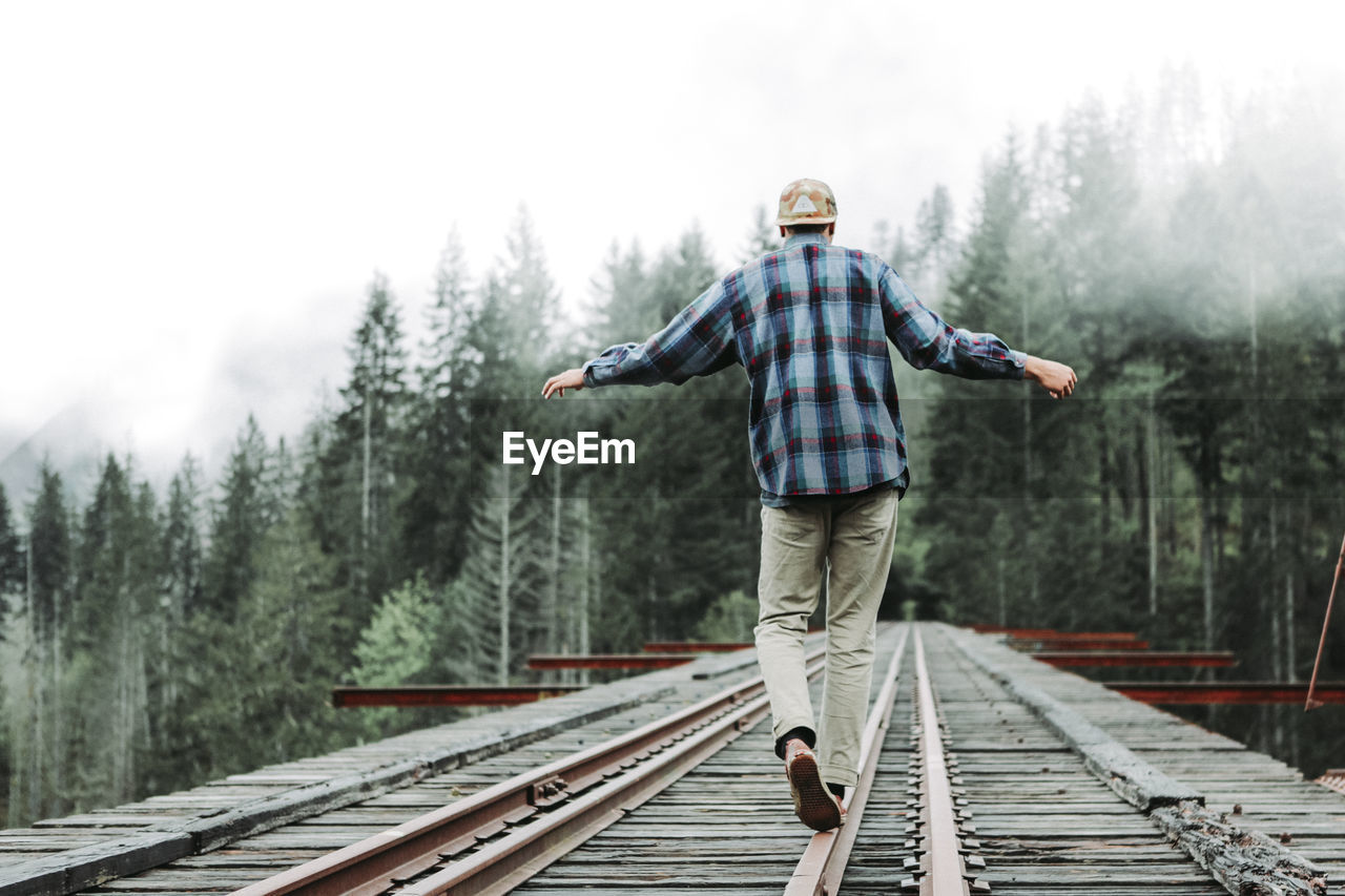 Young man balancing on railroad tracks over bridge in foggy forest
