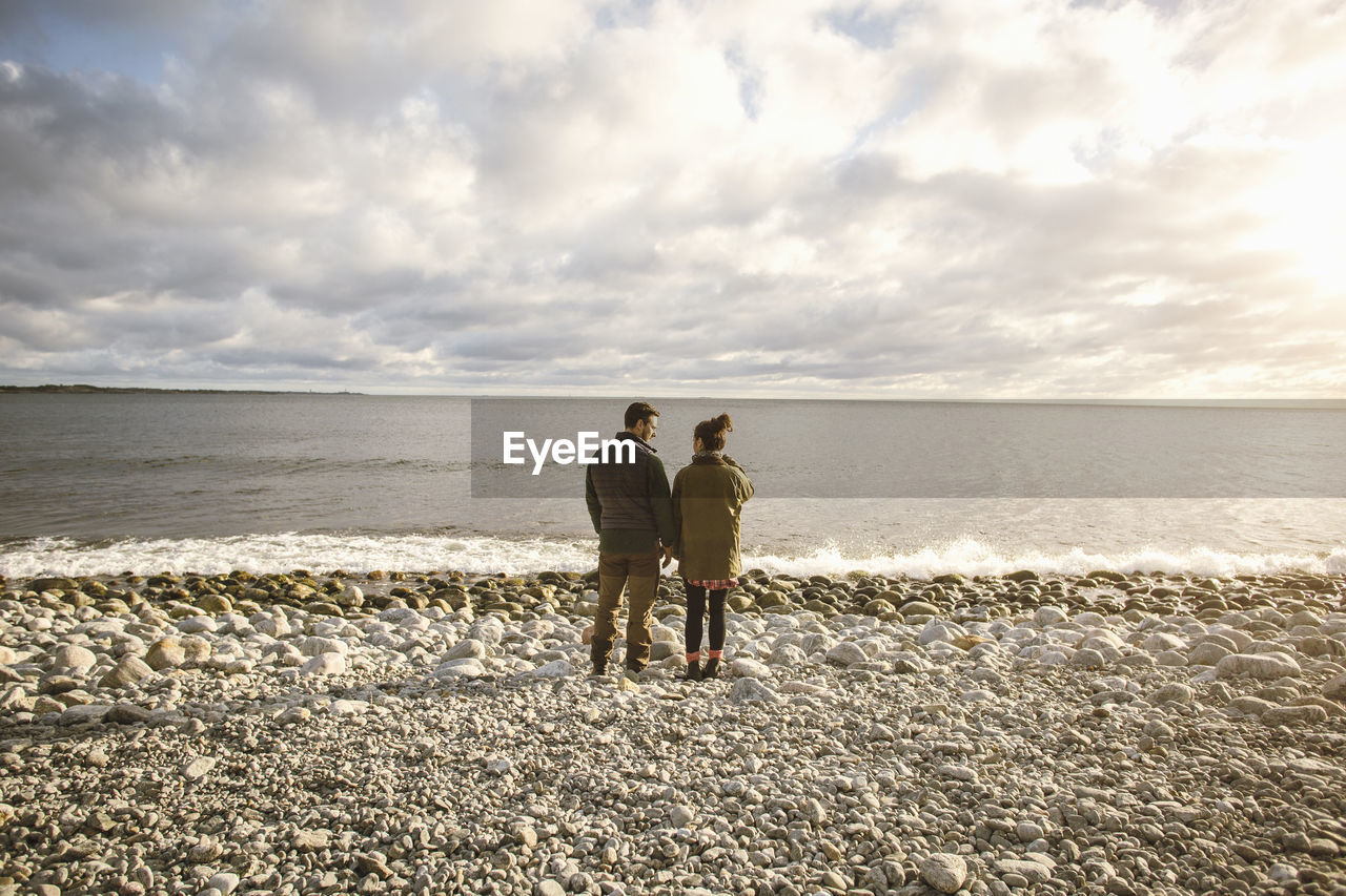 Rear view of couple standing on rocky shore at beach against sky