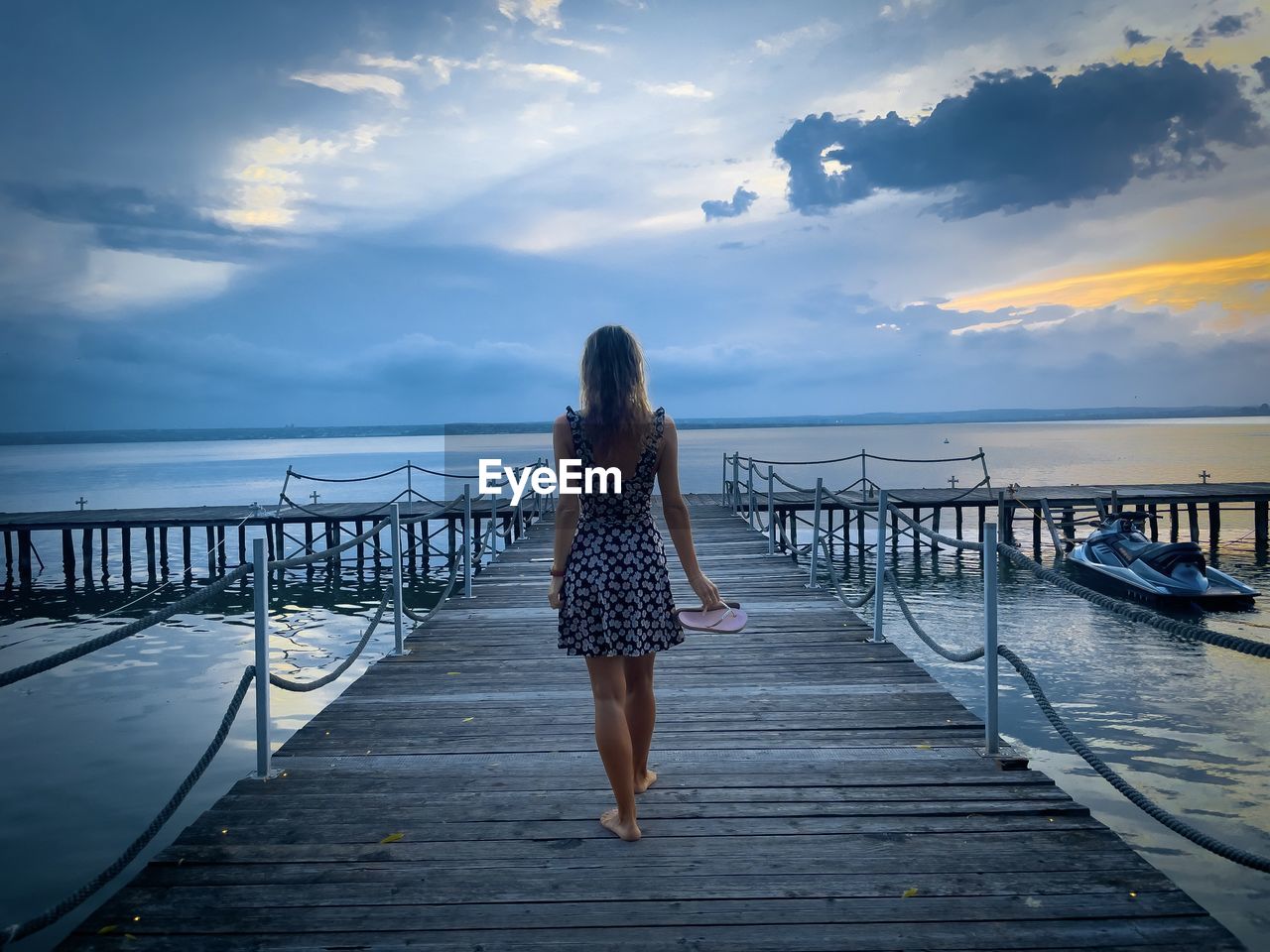 Woman in summer dress walking down a wooden pontoon towards the sea at sunset