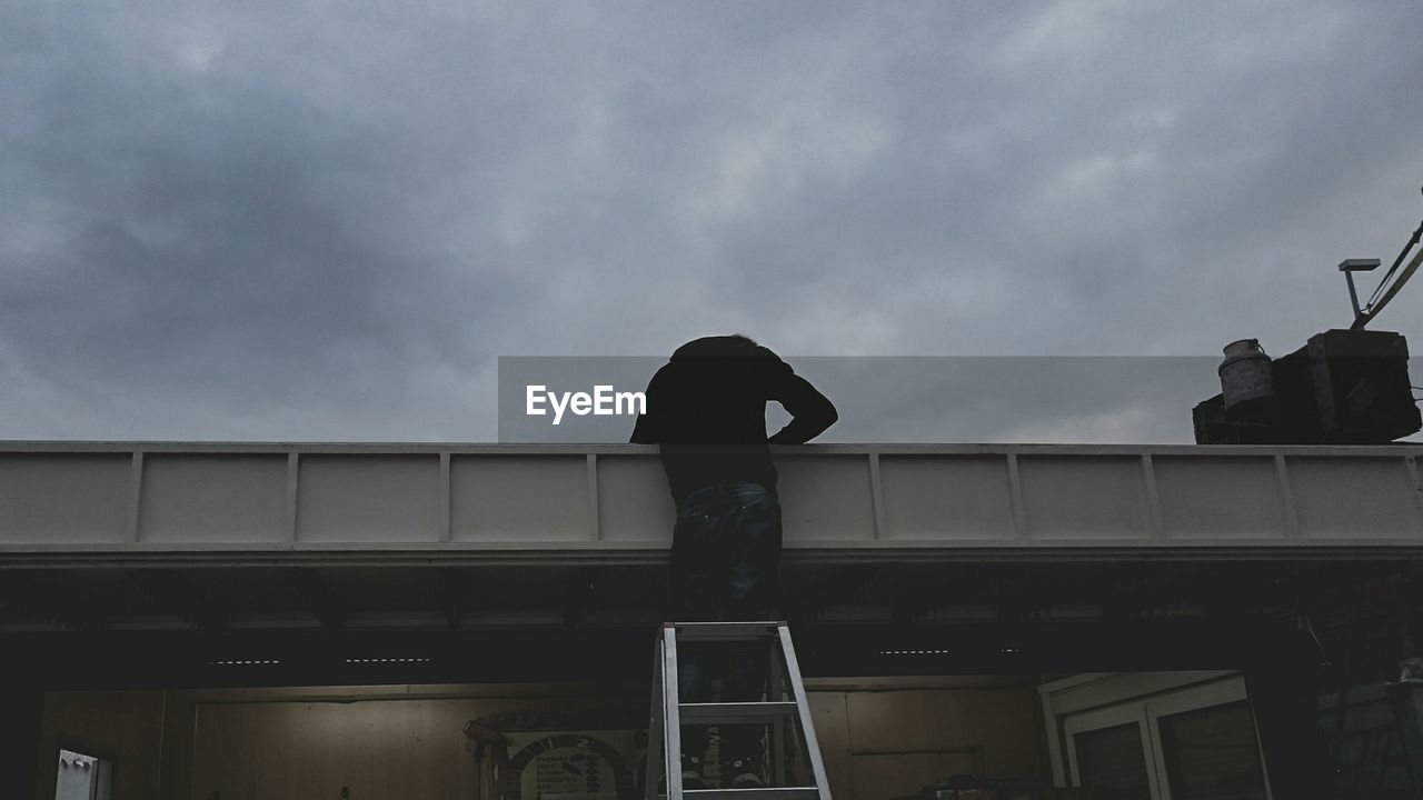 Rear view of man standing on ladder against cloudy sky