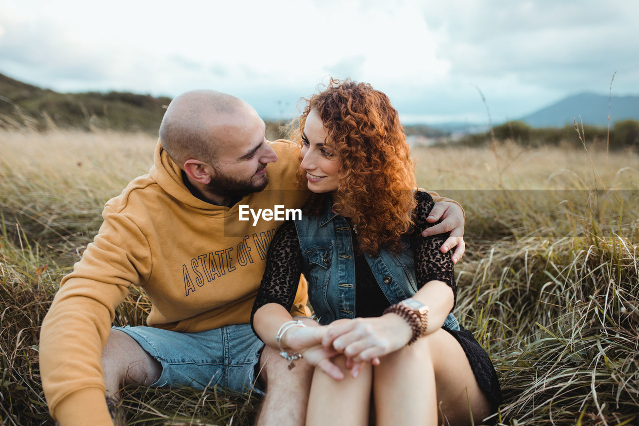 Young bald bearded man in yellow hoodie and jeans shorts embracing happy curly haired girlfriend in dress and denim vest while sitting together on meadow and talking