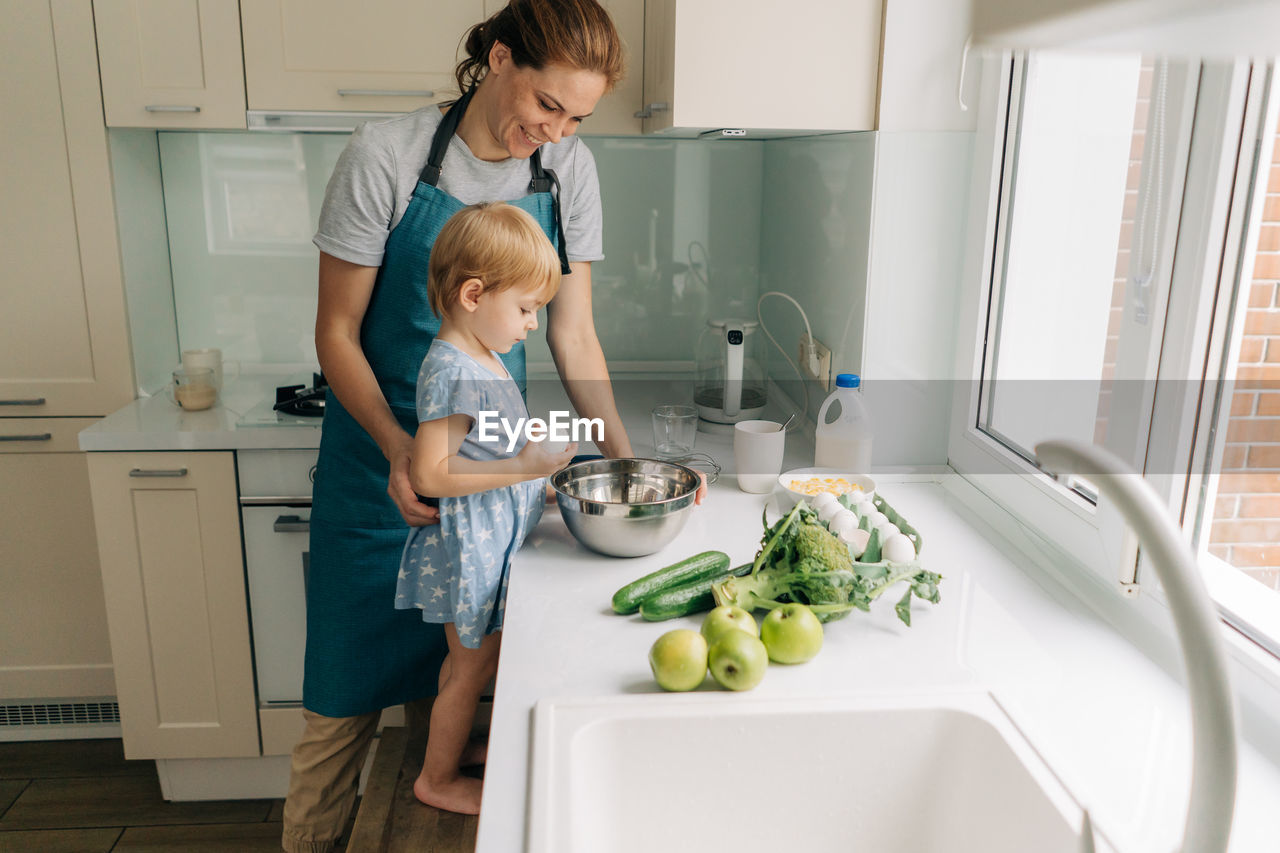 In a large bright kitchen a young mother and little daughter are preparing dinner.