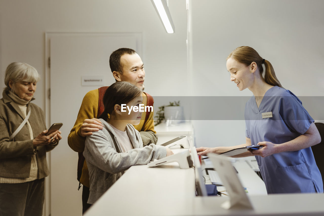 Smiling female healthcare worker assisting girl with father while booking appointment at hospital