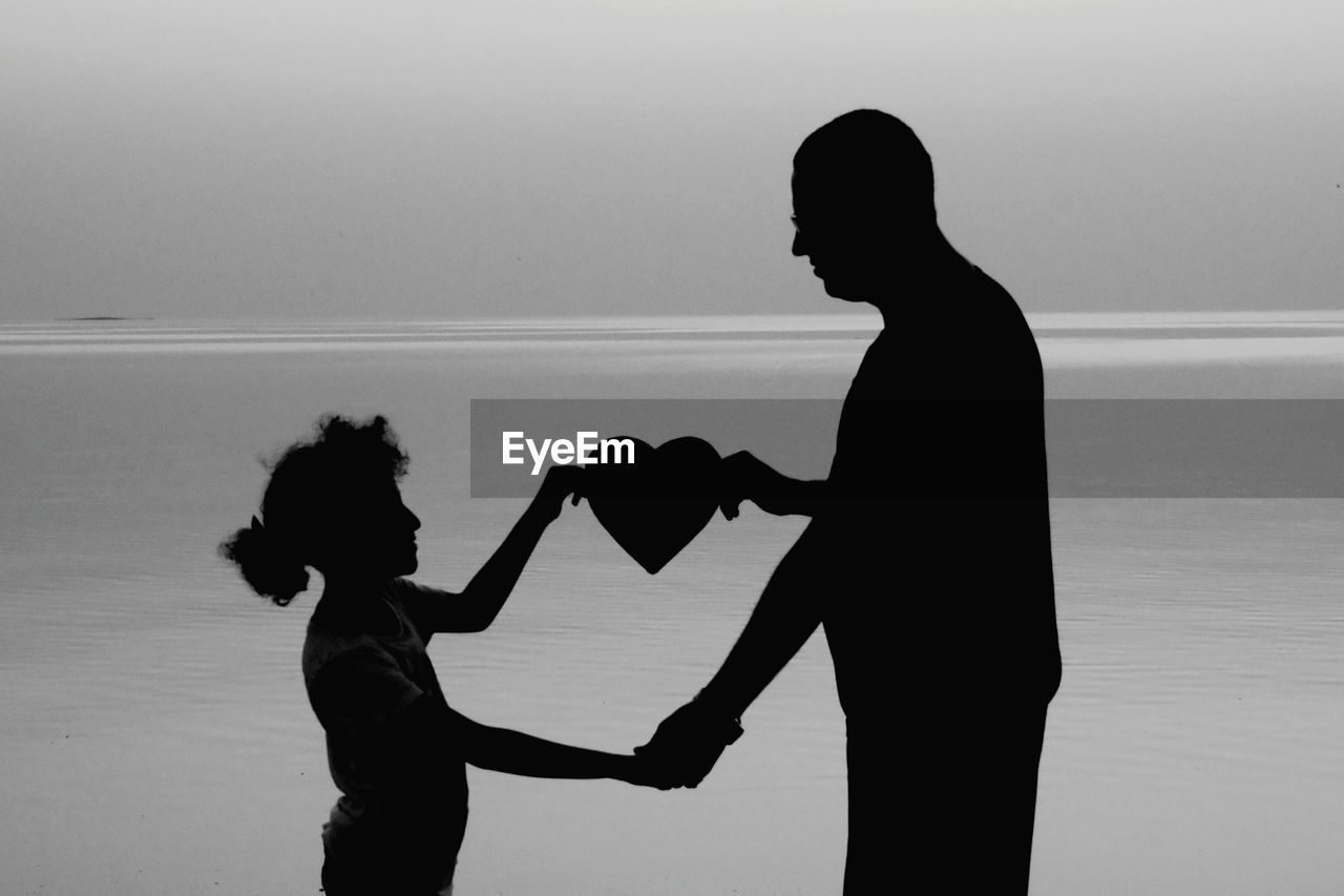 Silhouette father and daughter holding heart shape against sea