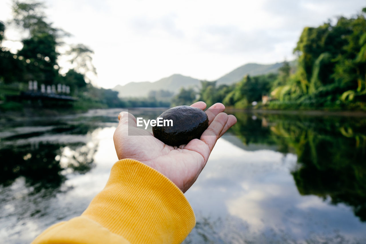 Close-up of person holding pebble over lake against sky