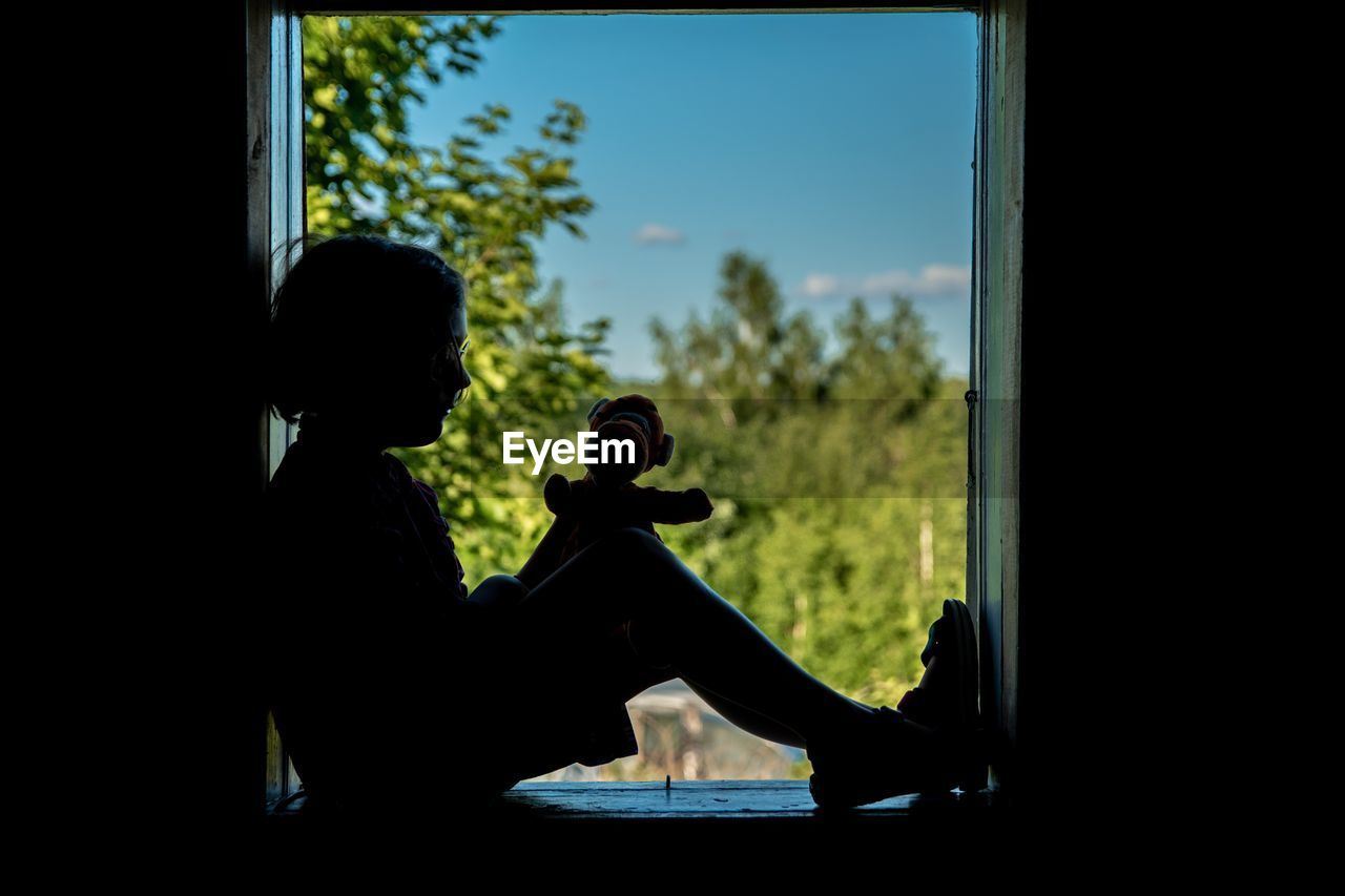 Side view of silhouette boy with toy sitting by window against sky