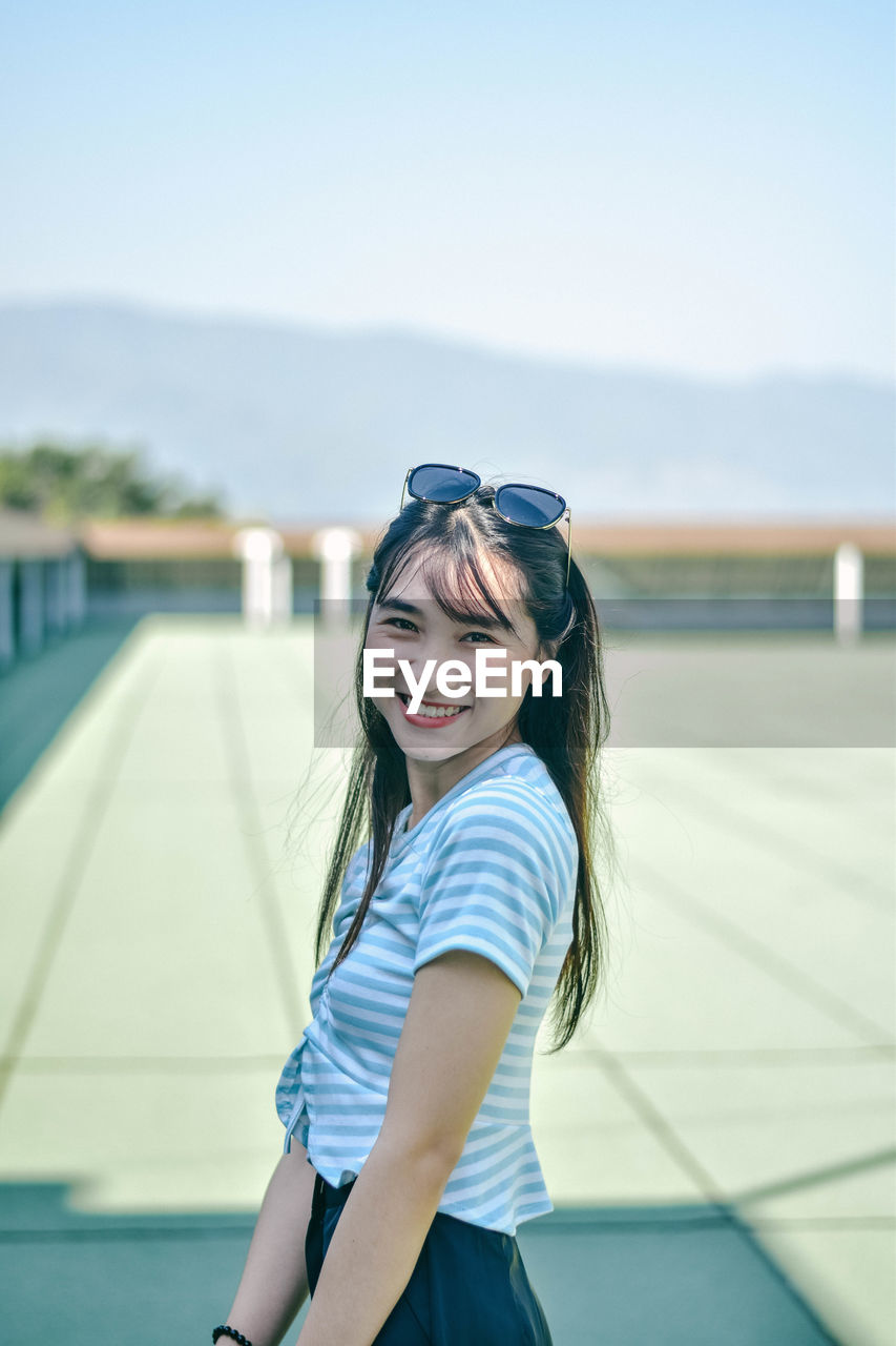 Portrait of smiling young woman standing at building terrace against sky