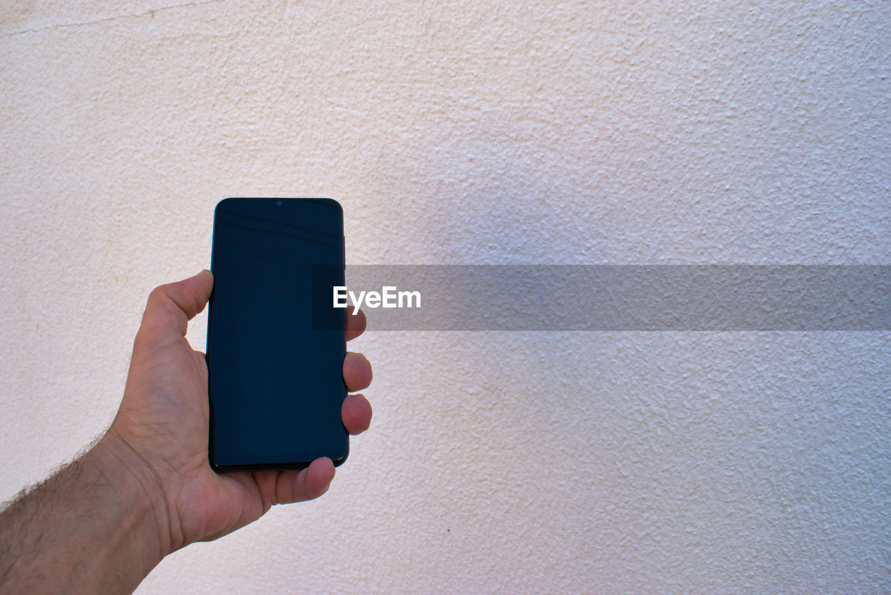 CLOSE-UP OF PERSON HOLDING SMART PHONE AGAINST WALL