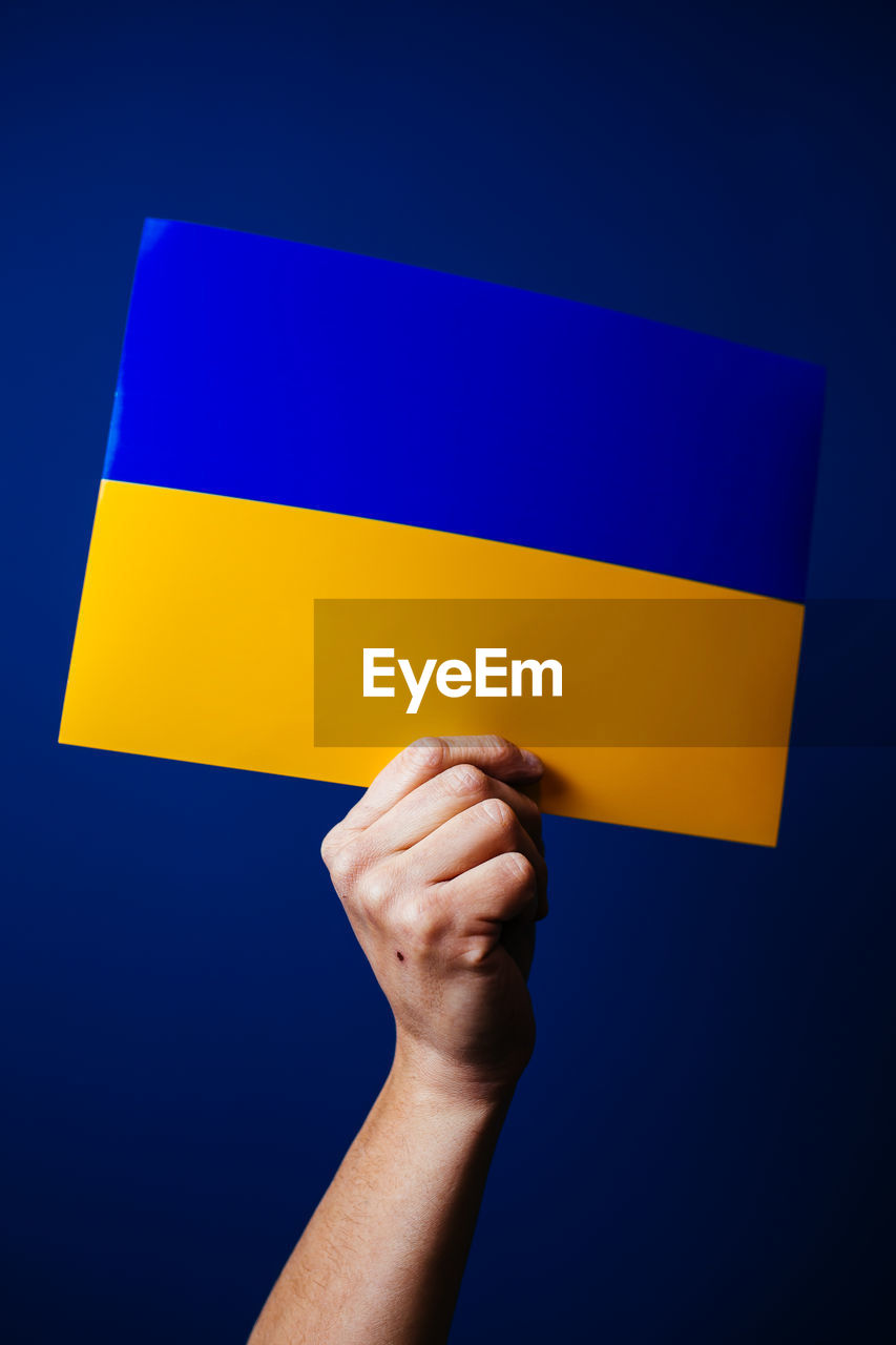 hand, blue, one person, holding, yellow, communication, studio shot, copy space, adult, symbol, sign, colored background, indoors, close-up, finger, flag, business, human limb, showing