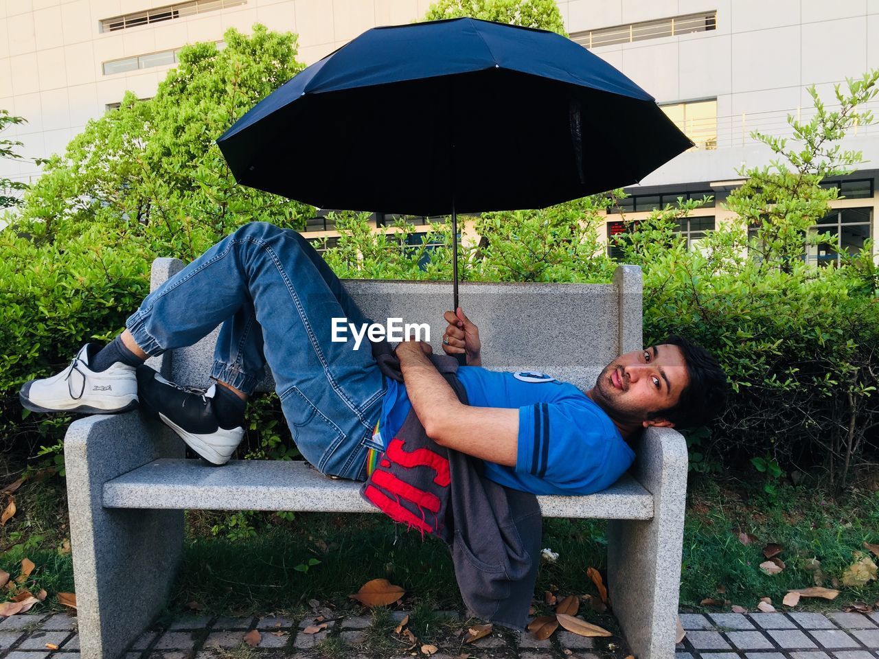 Portrait of young man holding umbrella while lying on bench at park