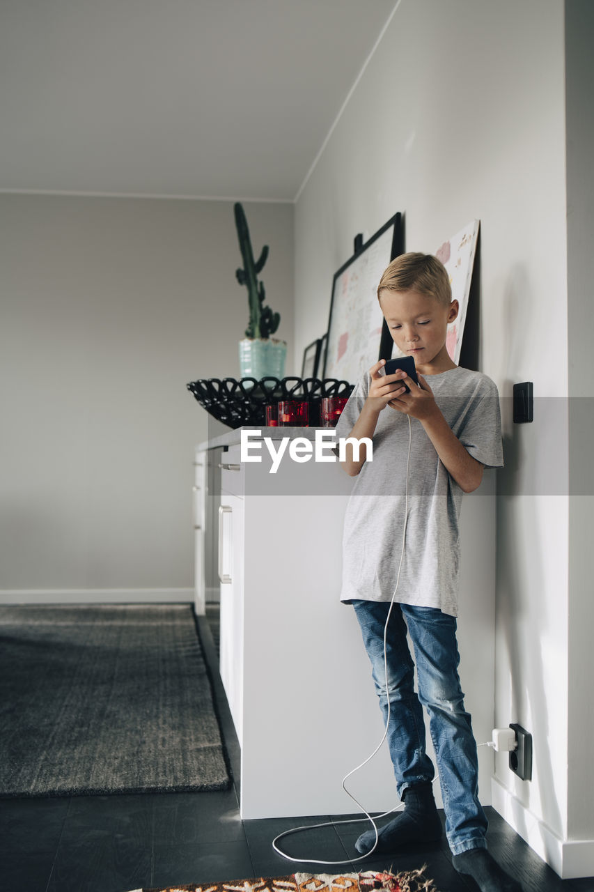 Full length of boy charging smart phone while standing by wall in room at home