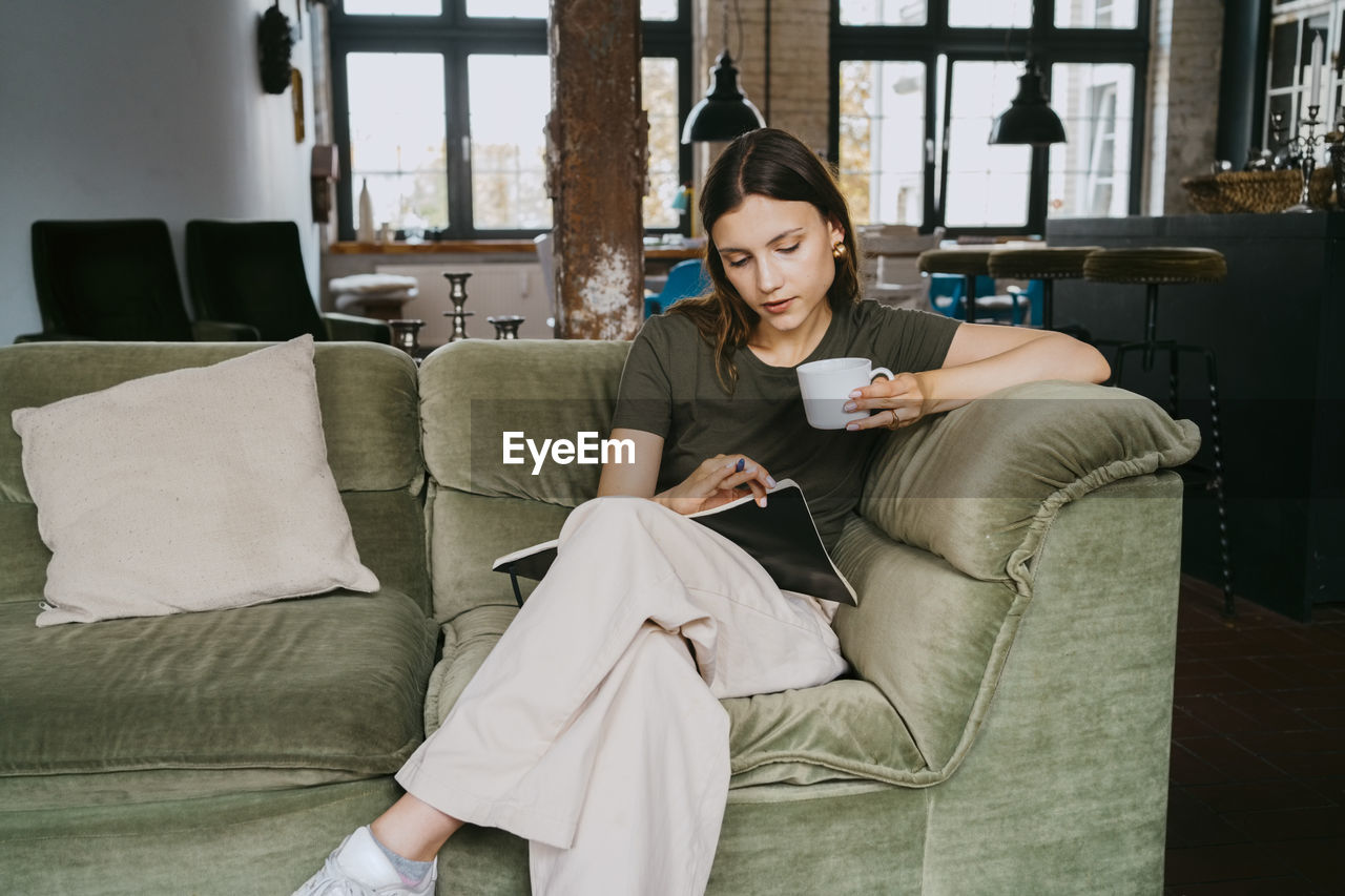 Young entrepreneur reading book having coffee while sitting on sofa in studio