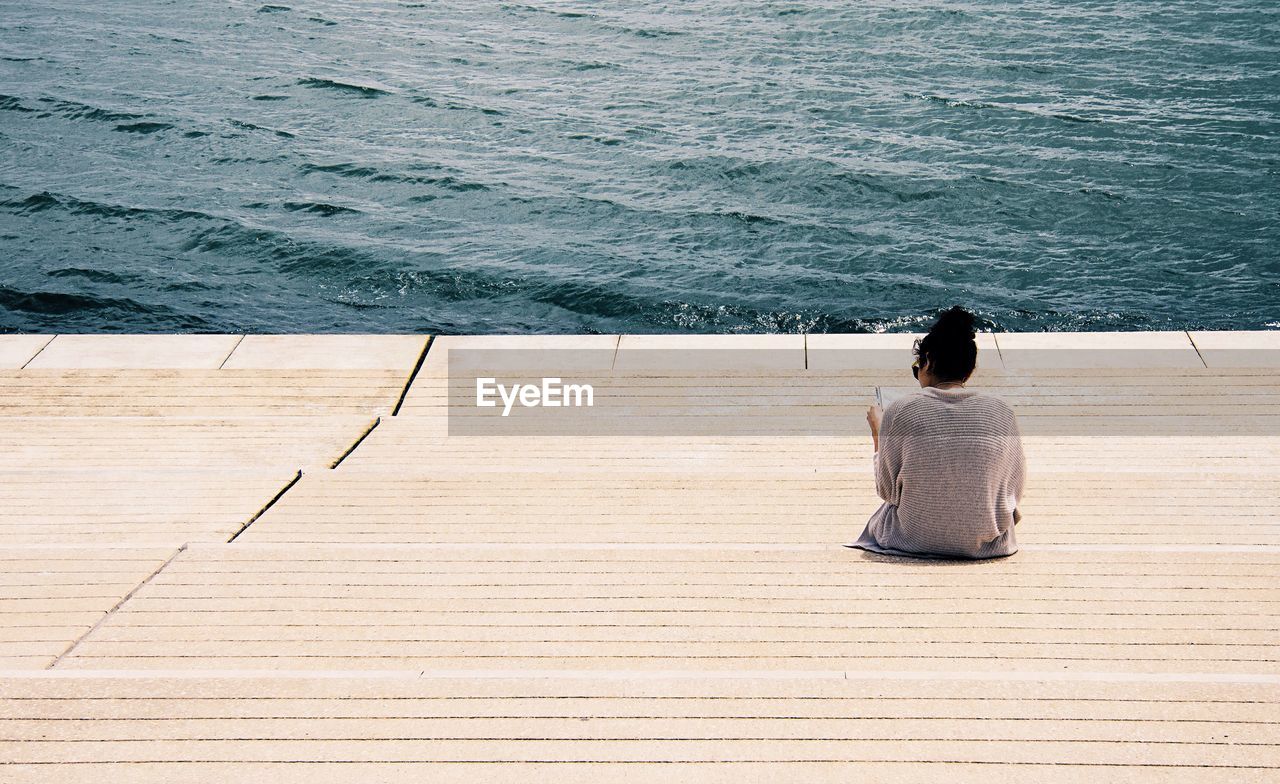 Rear view of woman sitting on steps against river during sunny day