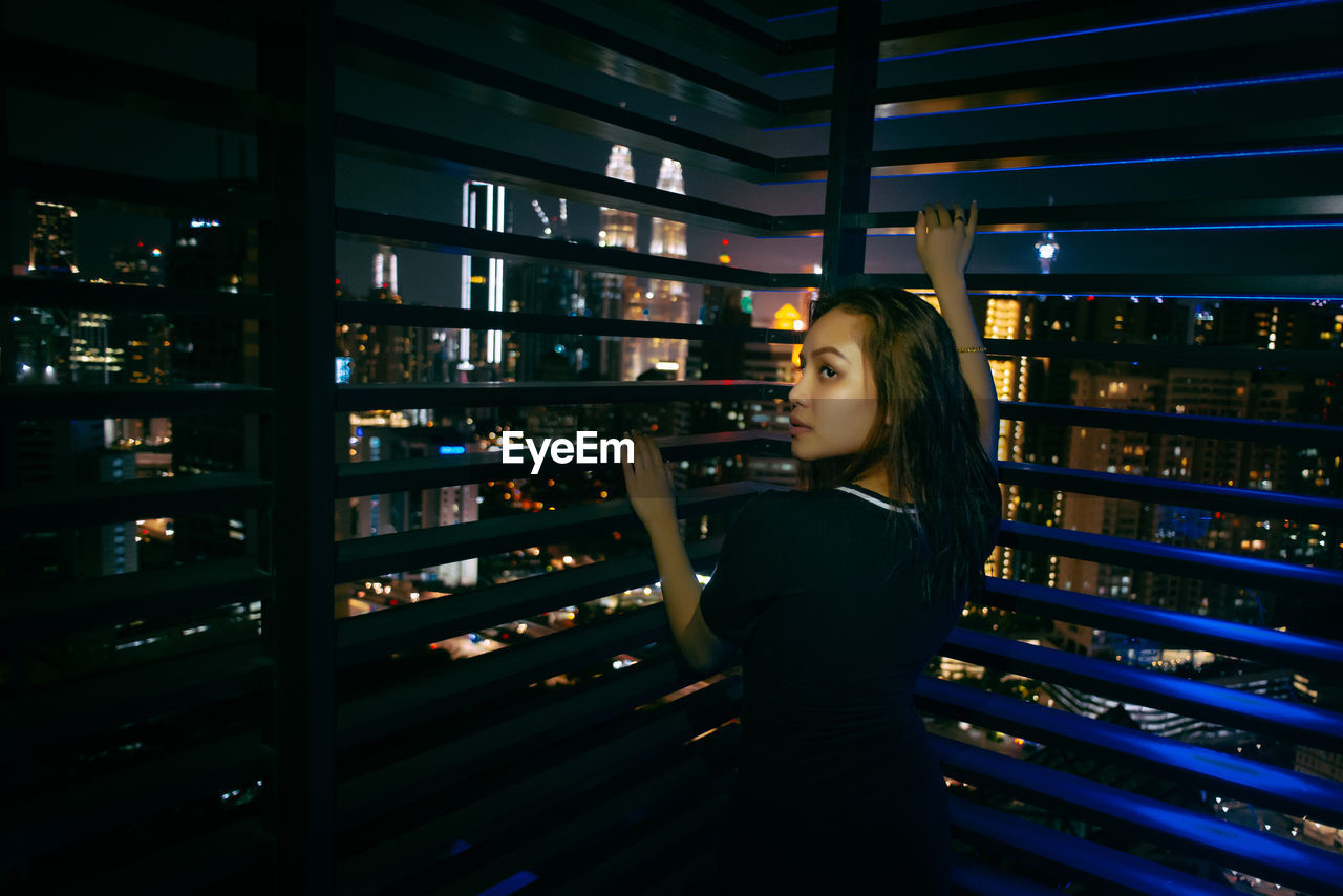 Young woman leaning on window blinds at night