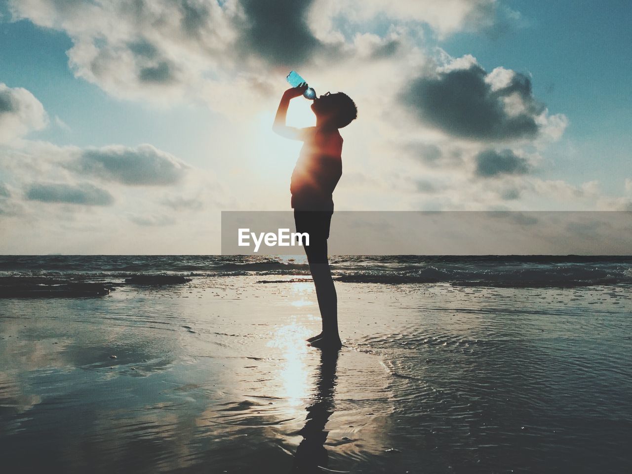 Side view of man standing and drinking water at sea shore against cloudy sky