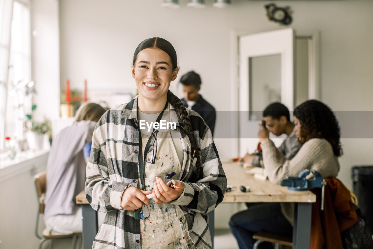 Portrait of smiling female teacher wearing plaid shirt with students in background at carpentry class