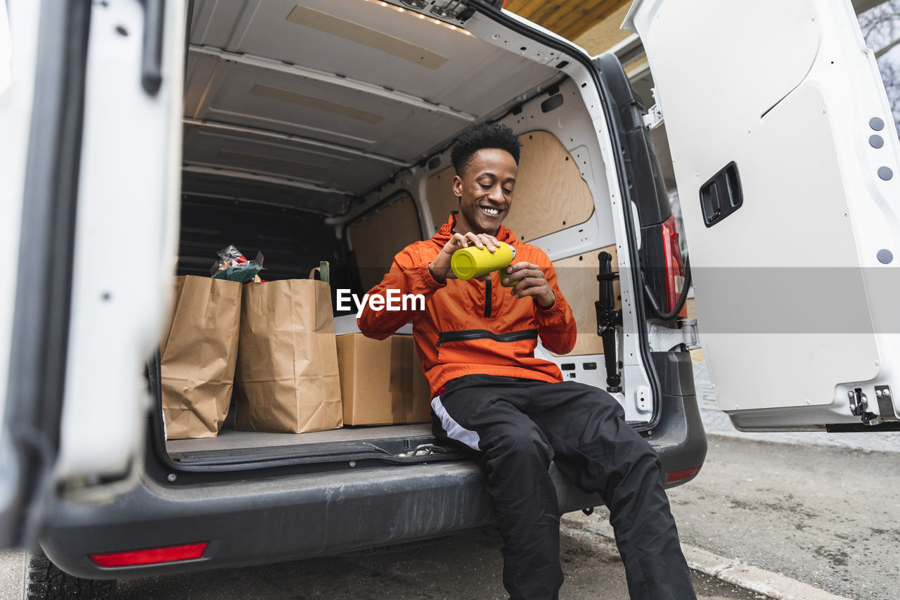 Happy male delivery person pouring coffee while sitting in van trunk