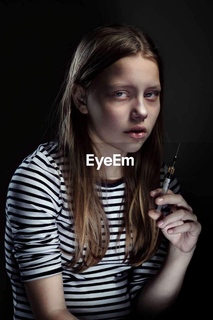 Portrait of young woman holding syringe against black background