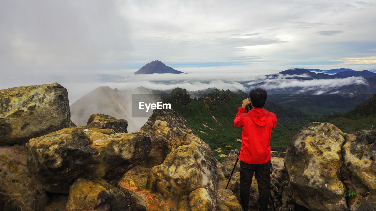 REAR VIEW OF MAN STANDING ON ROCKS AGAINST MOUNTAIN