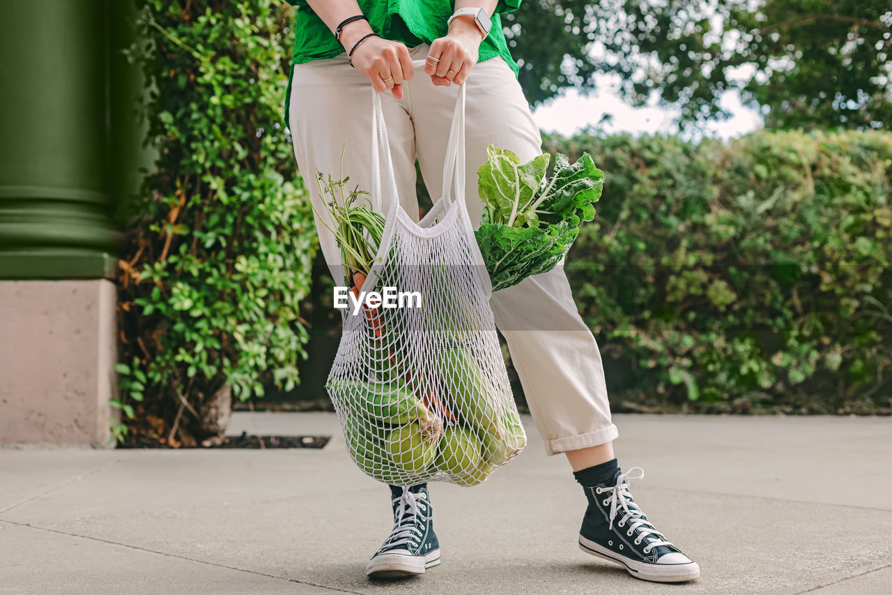 Unrecognizable woman in green shirt holding a mesh bag with veggies, standing on the street