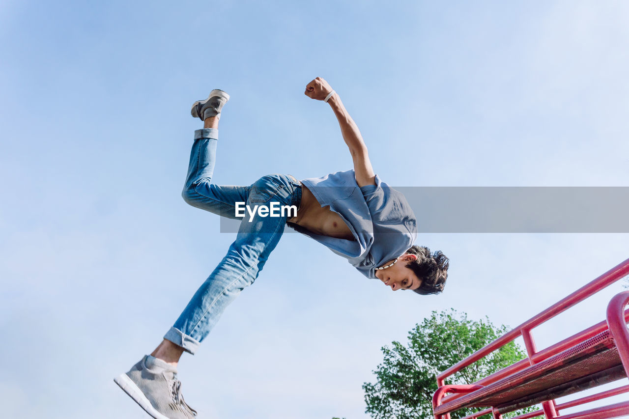 Low angle fearless young man jumping above metal railing in city while performing parkour stunt on sunny day