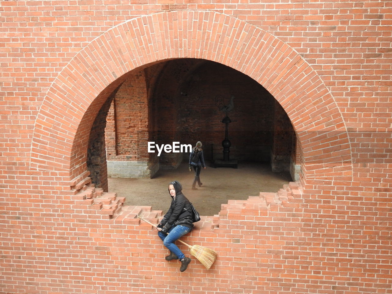 Side view of girl with broom sitting on brick wall