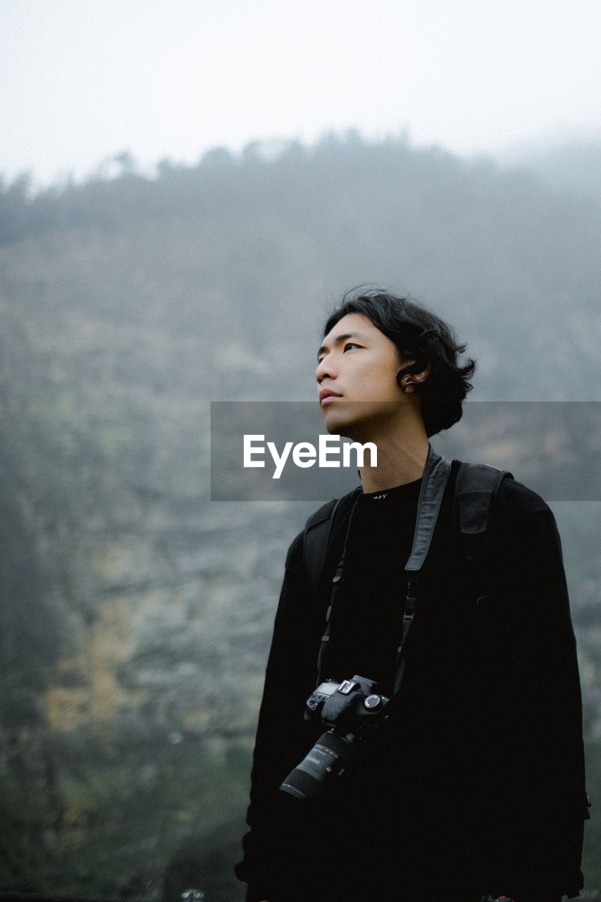 YOUNG WOMAN LOOKING AWAY WHILE STANDING IN FOGGY WEATHER