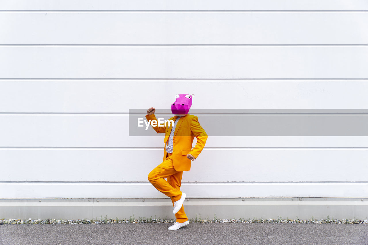 Man wearing vibrant orange suit and hippo mask jogging in front of white wall