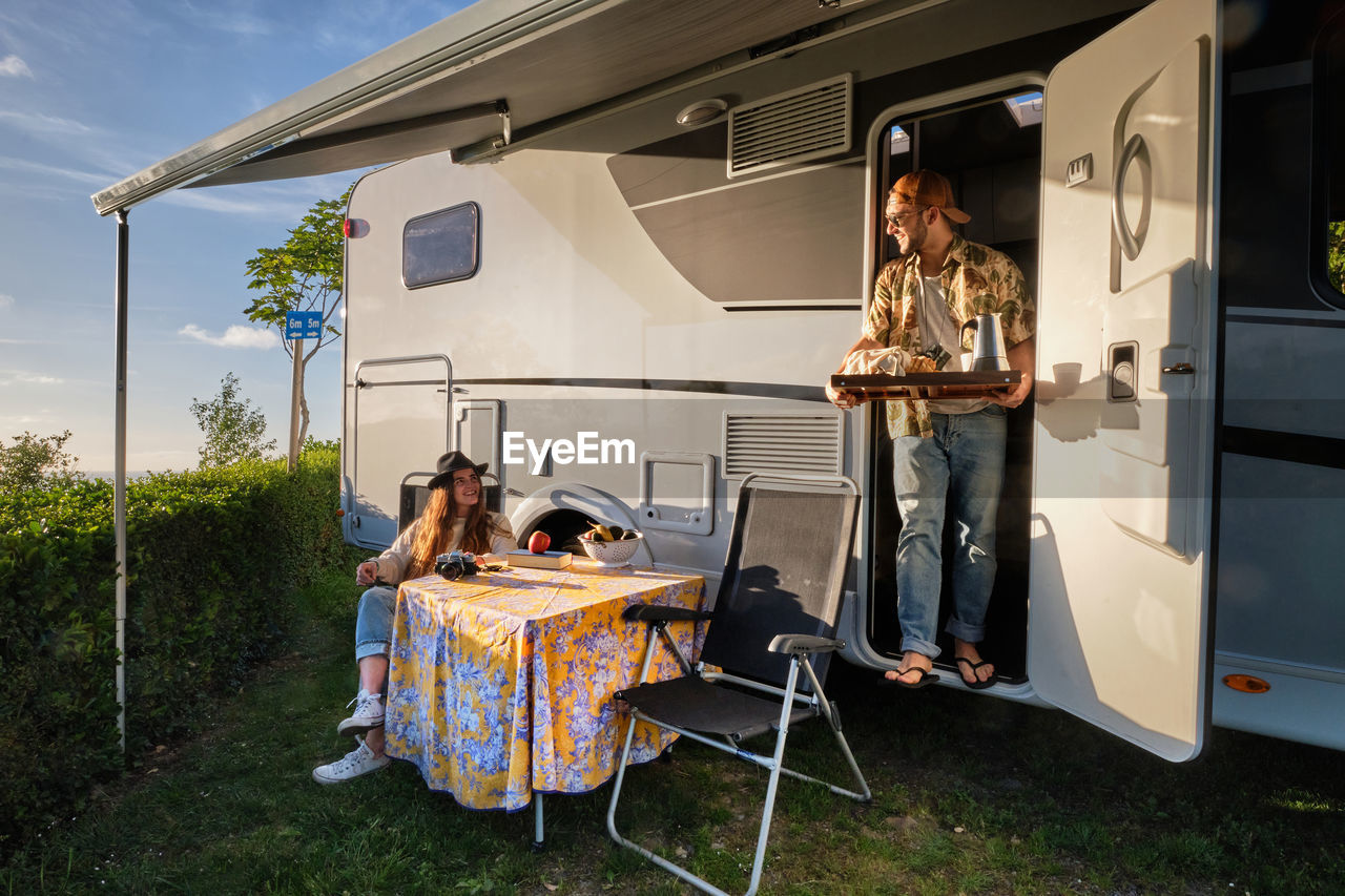 Cheerful traveling couple of hipsters having dinner at table near caravan during trip in summer in nature