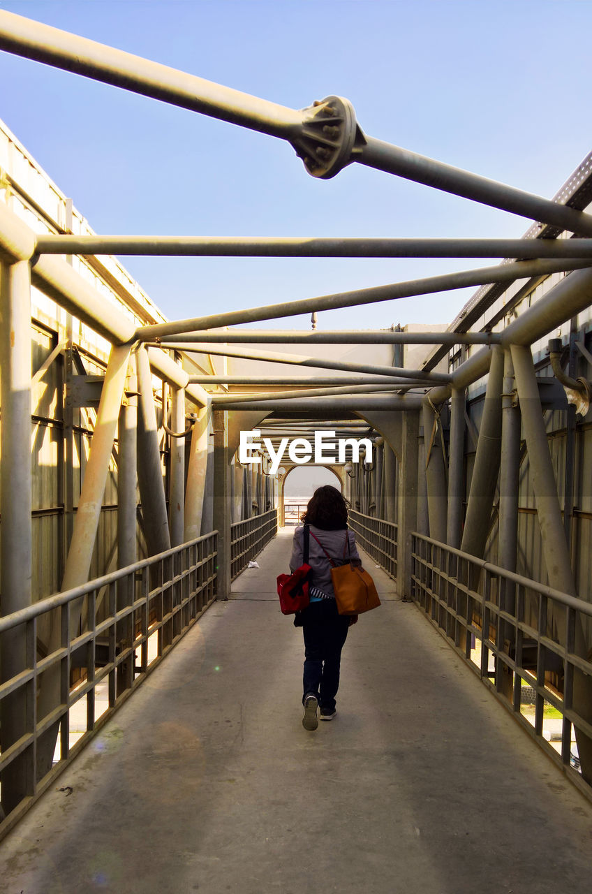 REAR VIEW OF WOMAN WALKING ON FOOTBRIDGE AGAINST SKY