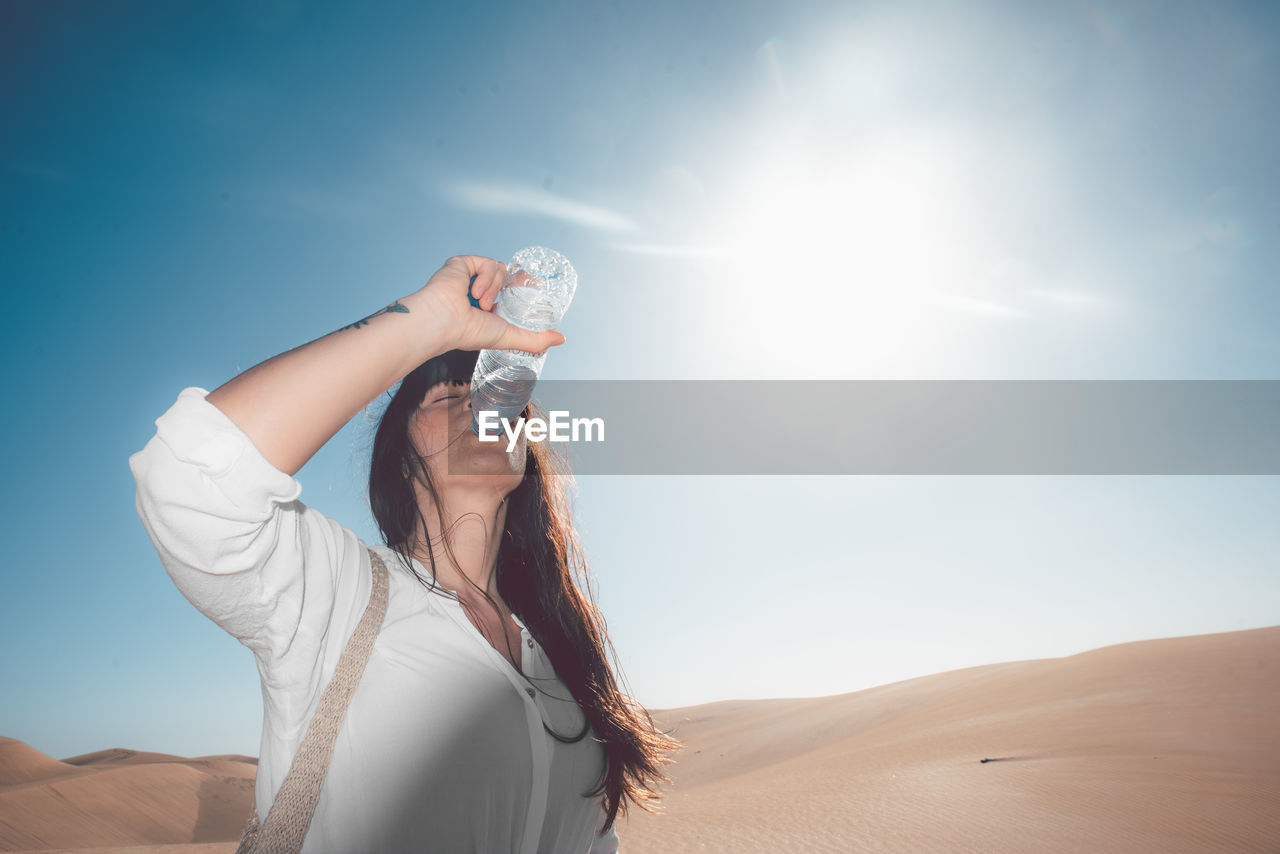 Low angle view of mid adult woman drinking water while standing at desert against sky during sunny day