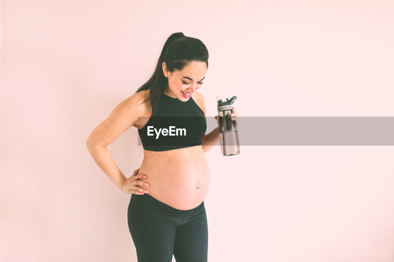 Smiling pregnant woman holding water bottle while standing against white wall