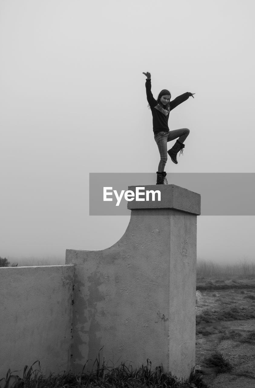 Low angle view of girl with arms outstretched standing on wall against clear sky