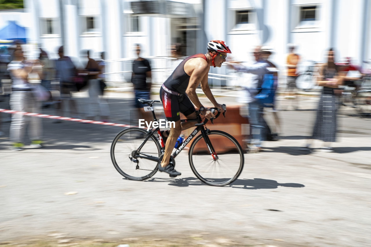 bicycle, transportation, sports, motion, activity, blurred motion, city, cycling, architecture, endurance sports, adult, road cycling, city life, vehicle, street, lifestyles, speed, men, road bicycle, city street, mode of transportation, sports equipment, commuter, racing bicycle, riding, wheel, building exterior, bicycle racing, exercising, clothing, person, full length, built structure, one person, on the move, cycle sport, footwear, headwear, helmet, copy space, road, travel, competition, recreation, race, athlete, outdoors, muscular build, bicycle wheel, road bicycle racing