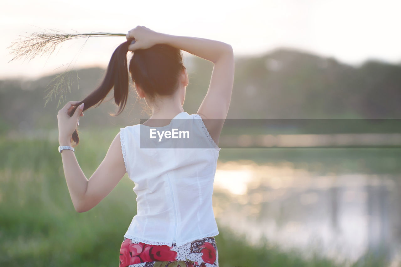 Rear view of woman tying up hair while standing on field during sunset