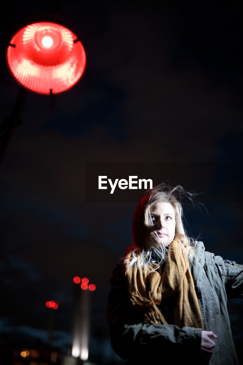 Portrait of young woman standing against illuminated city at night