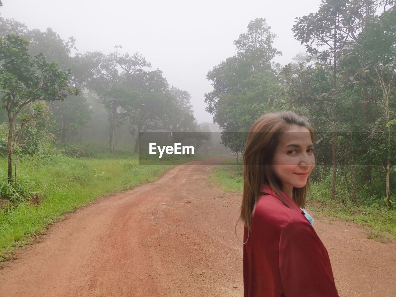 one person, tree, plant, dirt road, women, nature, portrait, road, land, rural area, fog, soil, environment, adult, young adult, landscape, lifestyles, forest, smiling, female, waist up, red, hairstyle, looking at camera, leisure activity, natural environment, dirt, outdoors, day, long hair, beauty in nature, clothing, non-urban scene, rural scene, casual clothing, child, tranquility, standing, sky, transportation, happiness