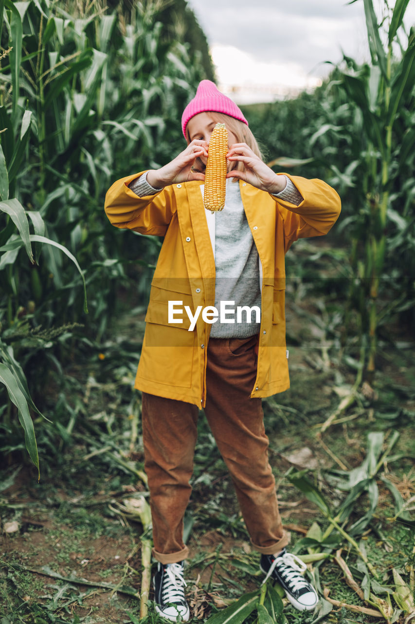 Funny little girl dressed in a yellow raincoat and a hot pink cap spoils and bites corn 