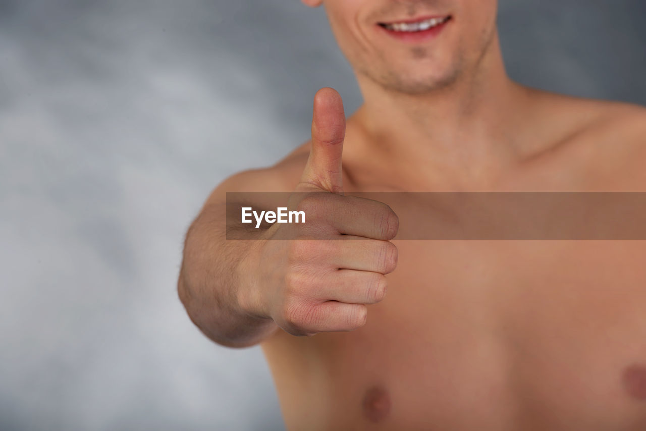 Midsection of shirtless man showing thumbs up sign against gray background