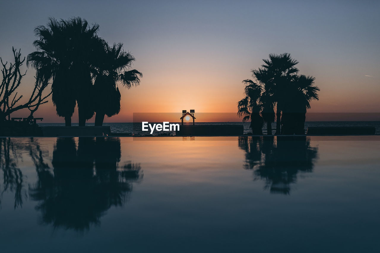 Silhouette palm trees by swimming pool against sky during sunset