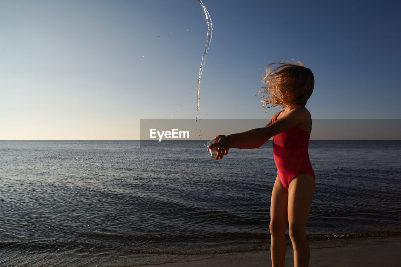 Cute girl throwing water at beach against sky