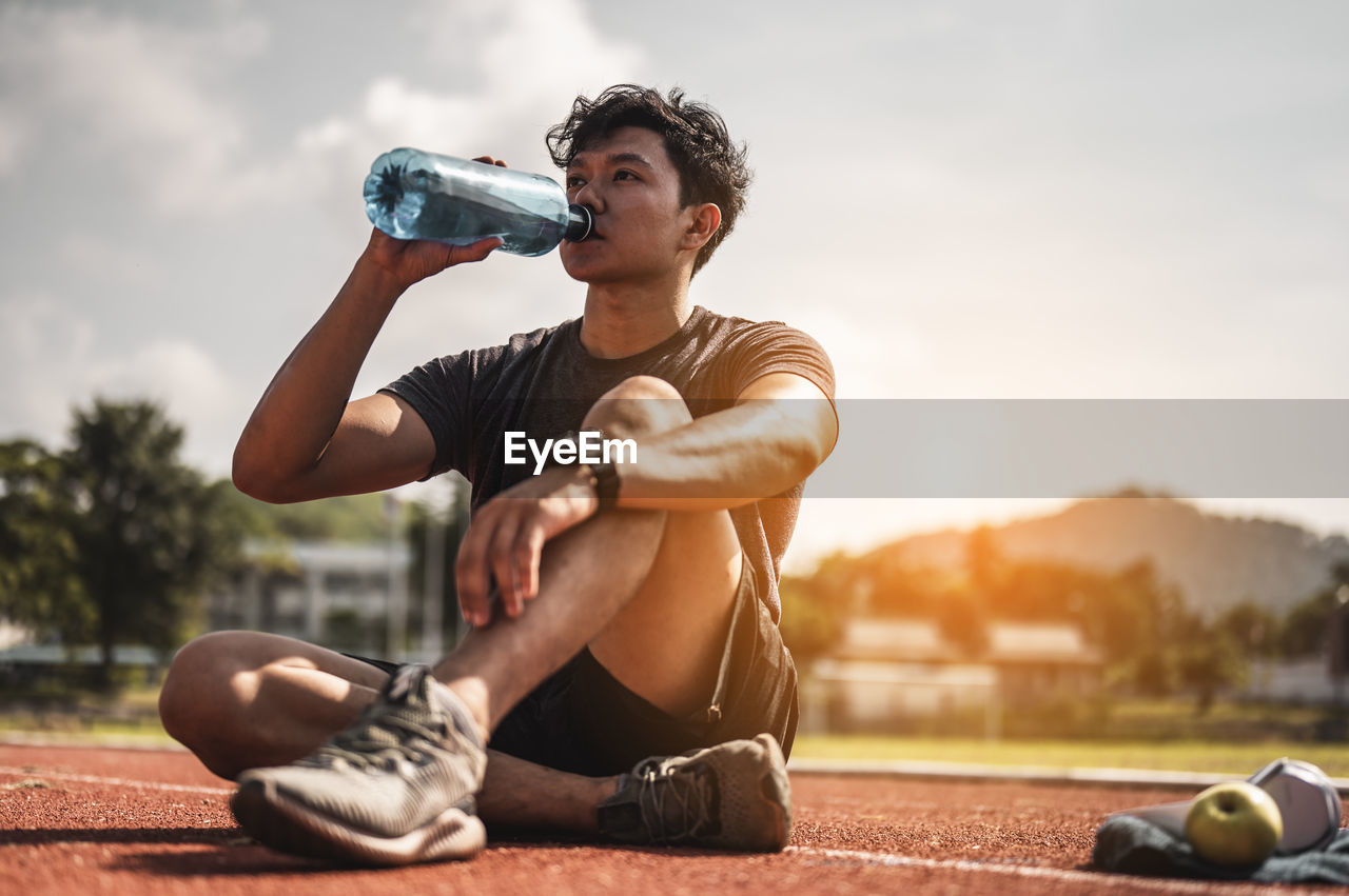 Full length of young man sitting at bottle against sky