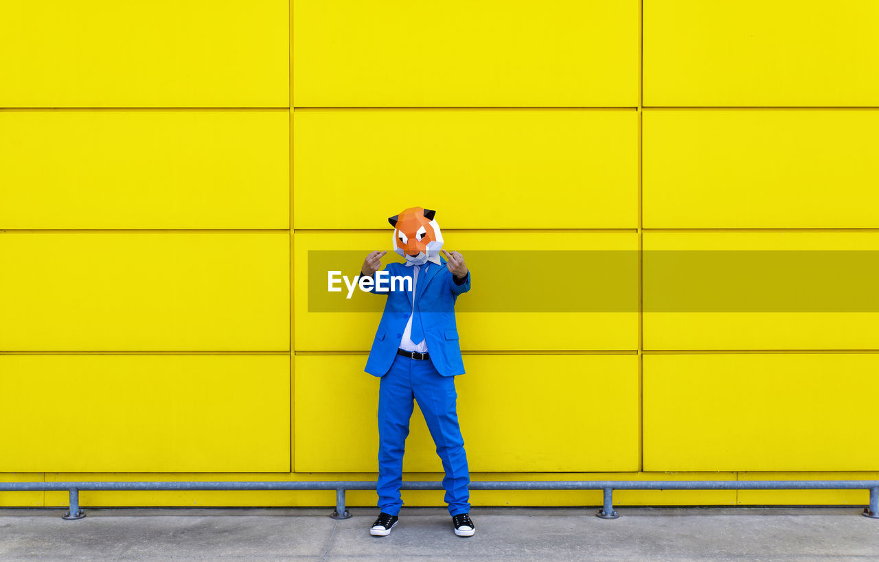 Man wearing vibrant blue suit and tiger mask showing two middle fingers in front of yellow wall