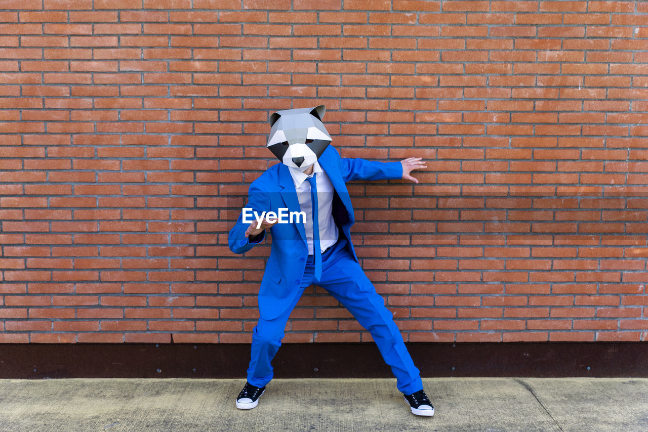 Man wearing vibrant blue suit and raccoon mask posing in front of brick wall